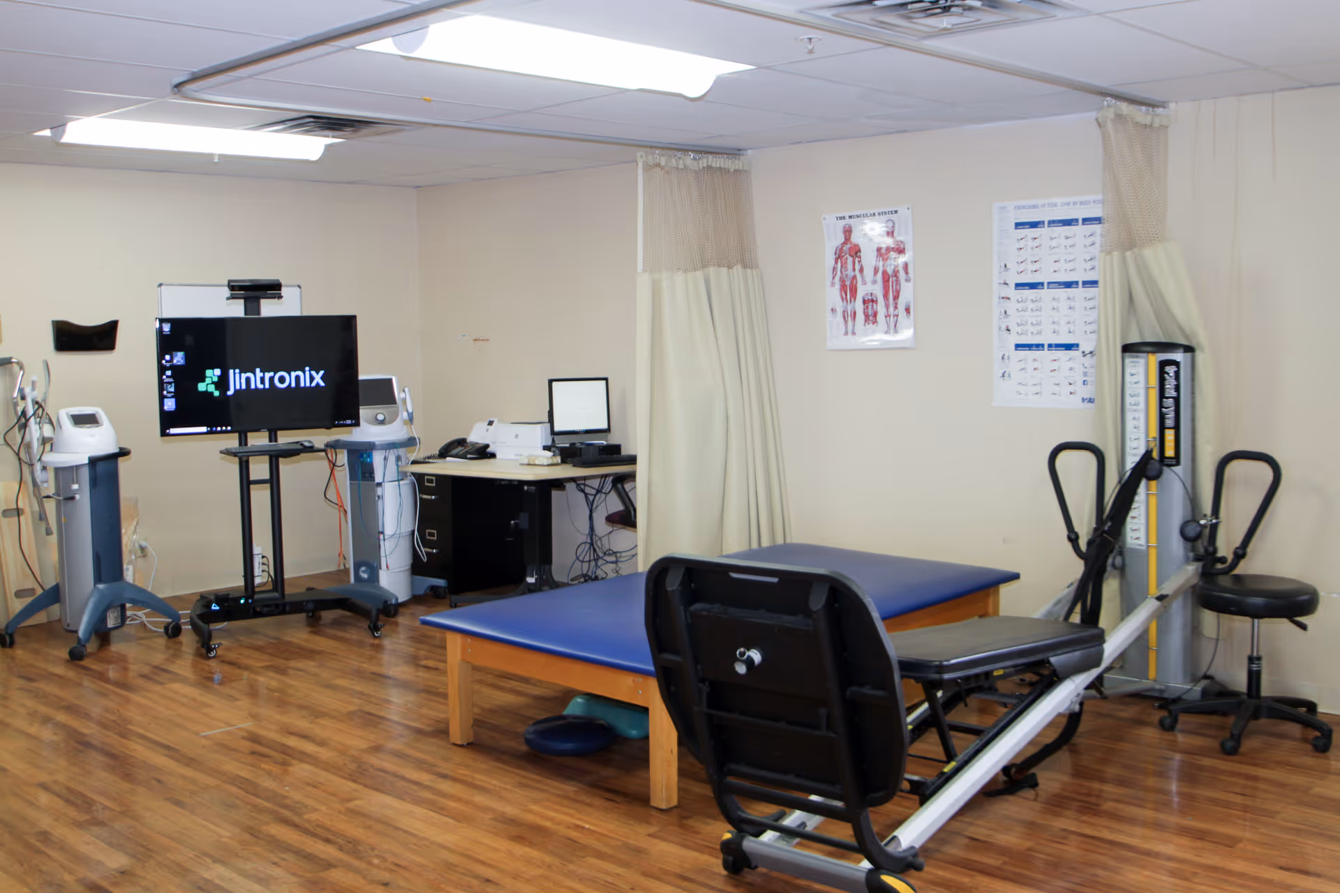 A rehabilitation therapy room with exercise equipment, a treatment table, medical devices and a monitor on wood flooring.