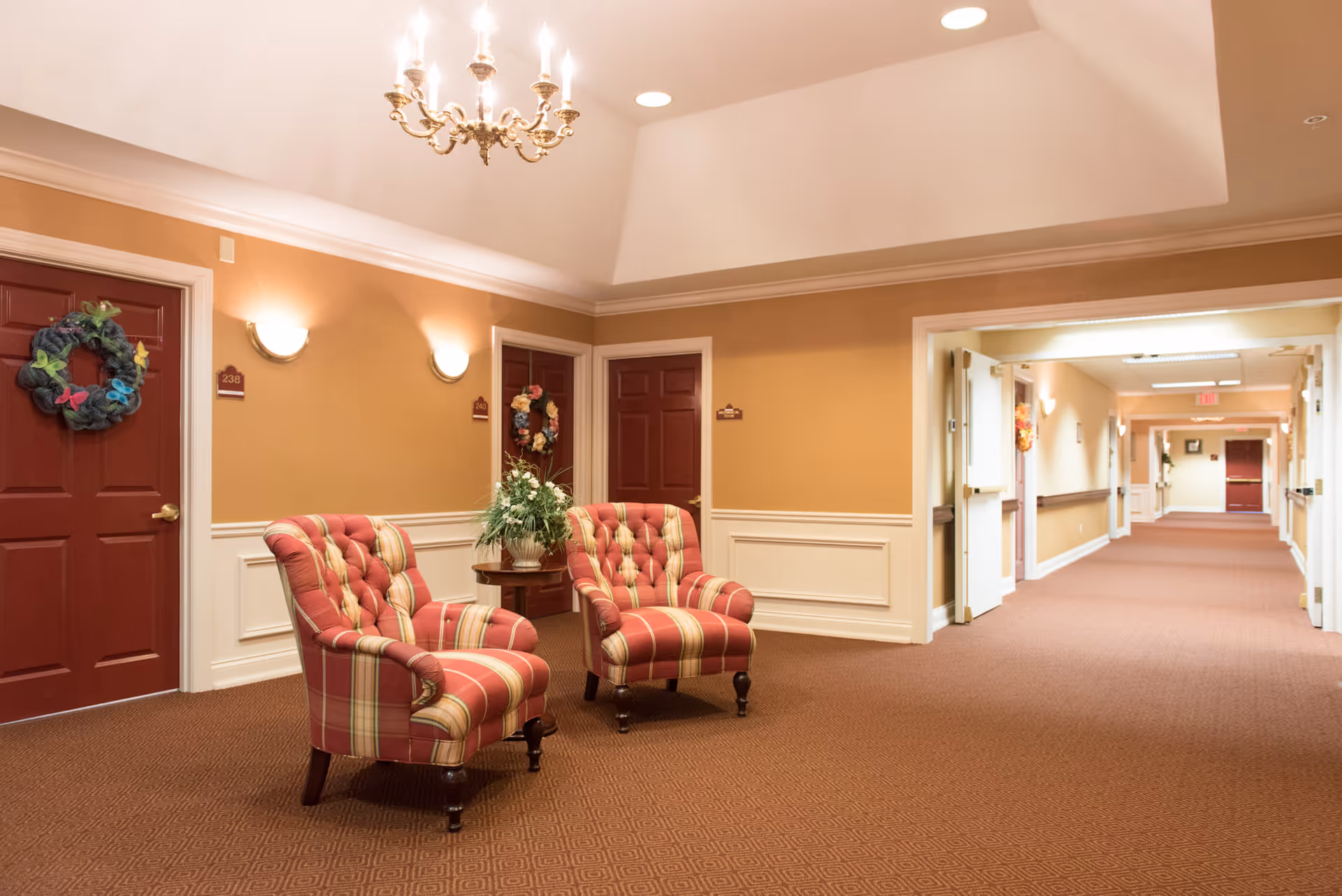 Carpeted interior hallway with two upholstered armchairs and a small table with a plant in front of red doors.