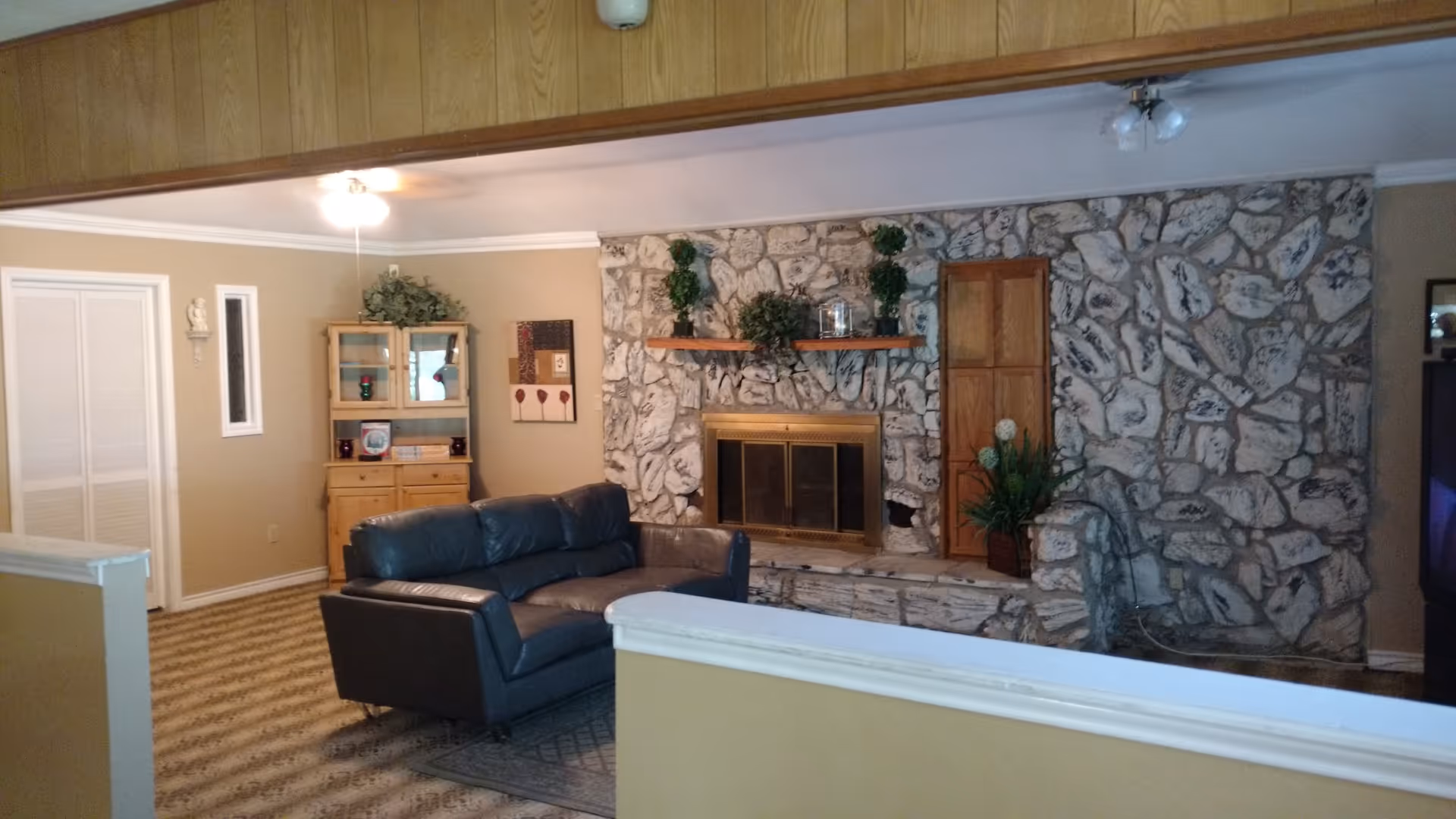 A living room with a stone fireplace as the focal point. The fireplace has a brass screen and a wooden shelf above it decorated with plants and a glass jar. To the left of the fireplace is a wooden cabinet with glass doors and decorative items on top and inside. A dark leather sofa is positioned in front of the fireplace on a patterned carpet. The walls are beige, and the ceiling has white crown molding with two ceiling lights visible.