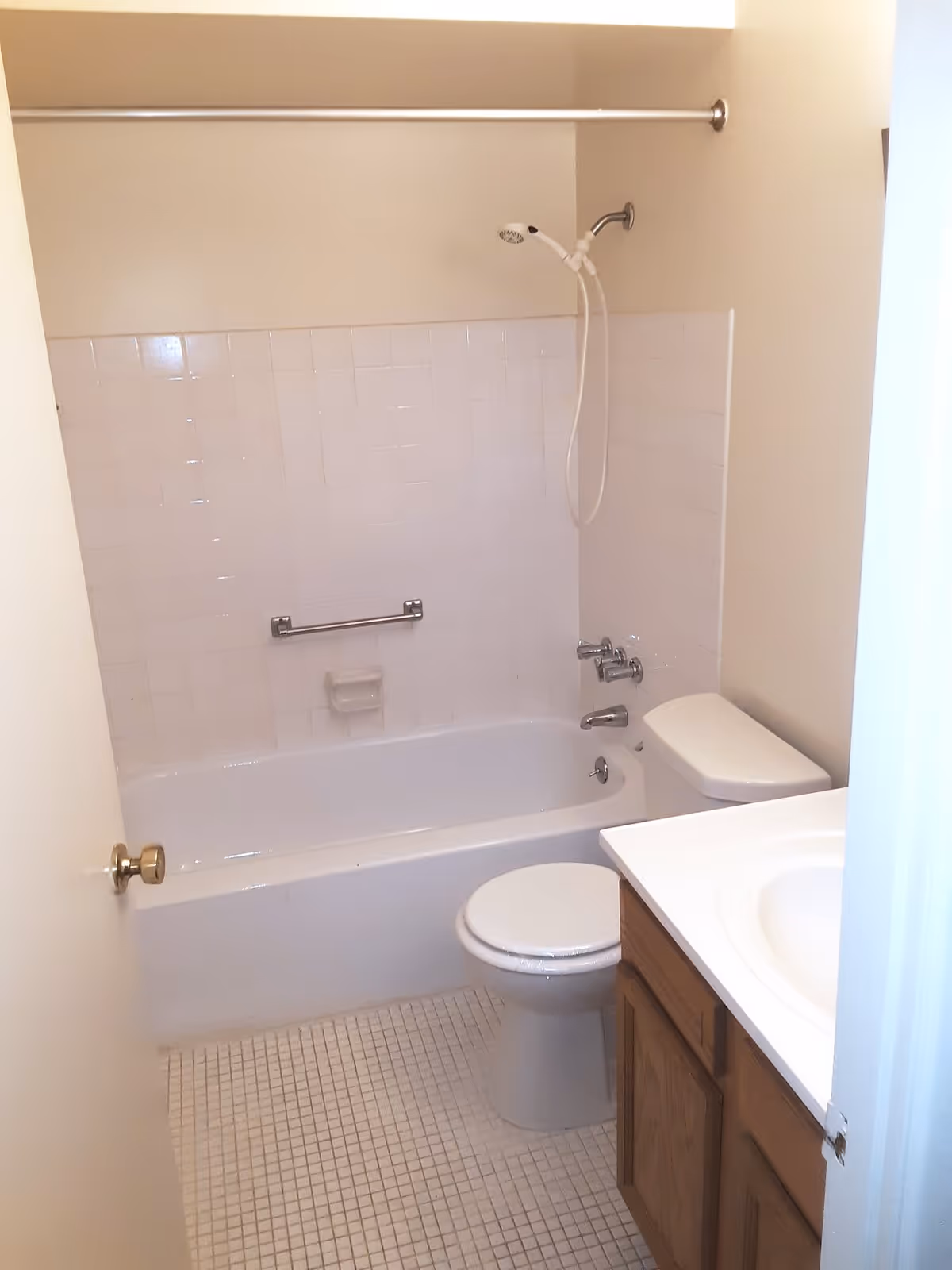 A small bathroom with a white bathtub and shower combination, a white toilet, a wooden vanity with a white sink, and beige tiled floor and walls.