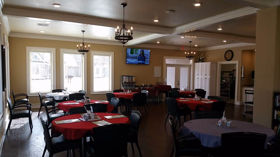 Dining room with round tables covered in red and purple tablecloths, black chairs, chandeliers, and a wall-mounted TV.