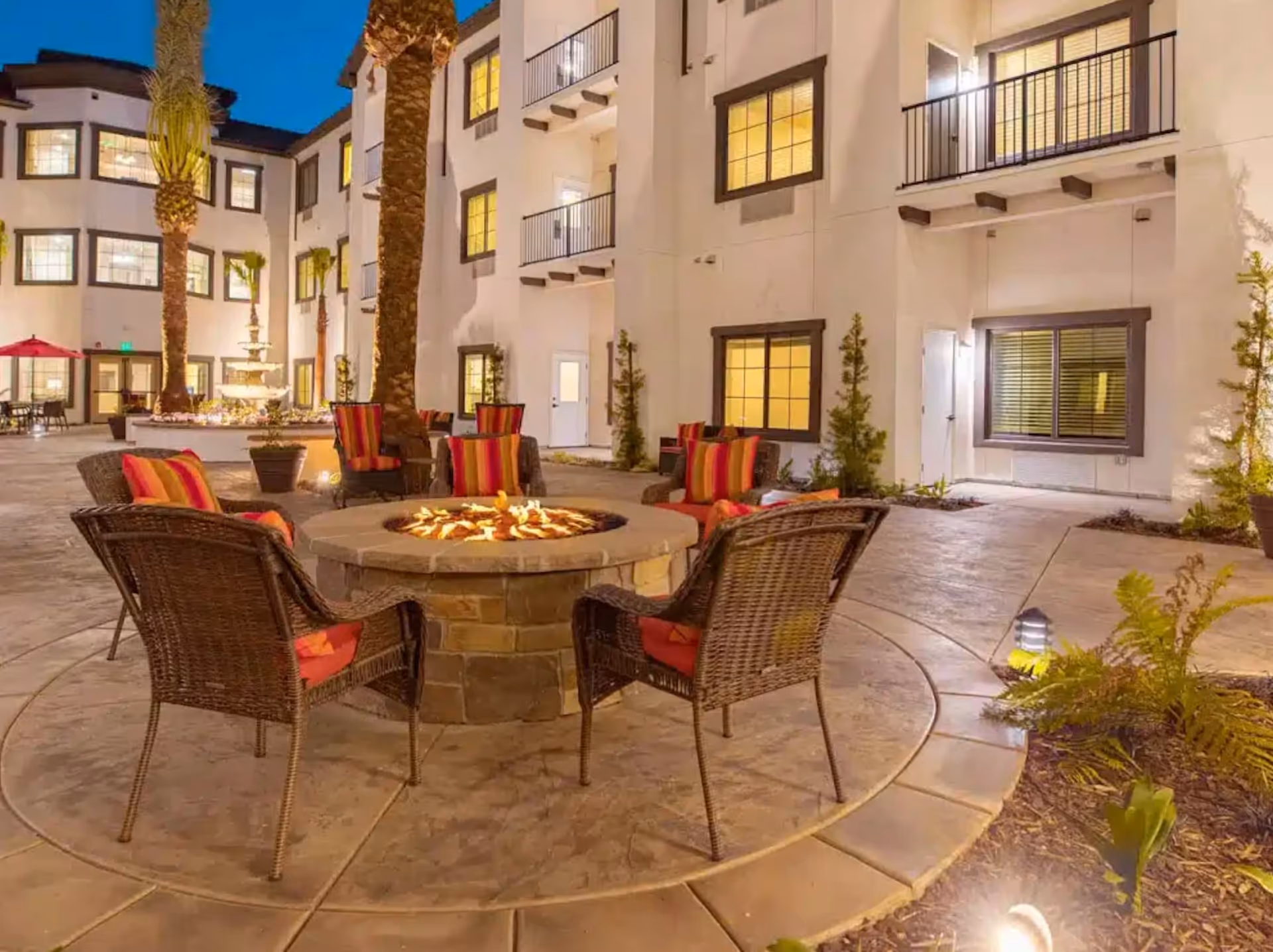 Outdoor courtyard at dusk with a circular stone fire pit surrounded by wicker chairs with red and orange striped cushions. The courtyard is part of a multi-story building with balconies and lit windows. There are palm trees and a water fountain in the background, along with some plants and pathway lighting.
