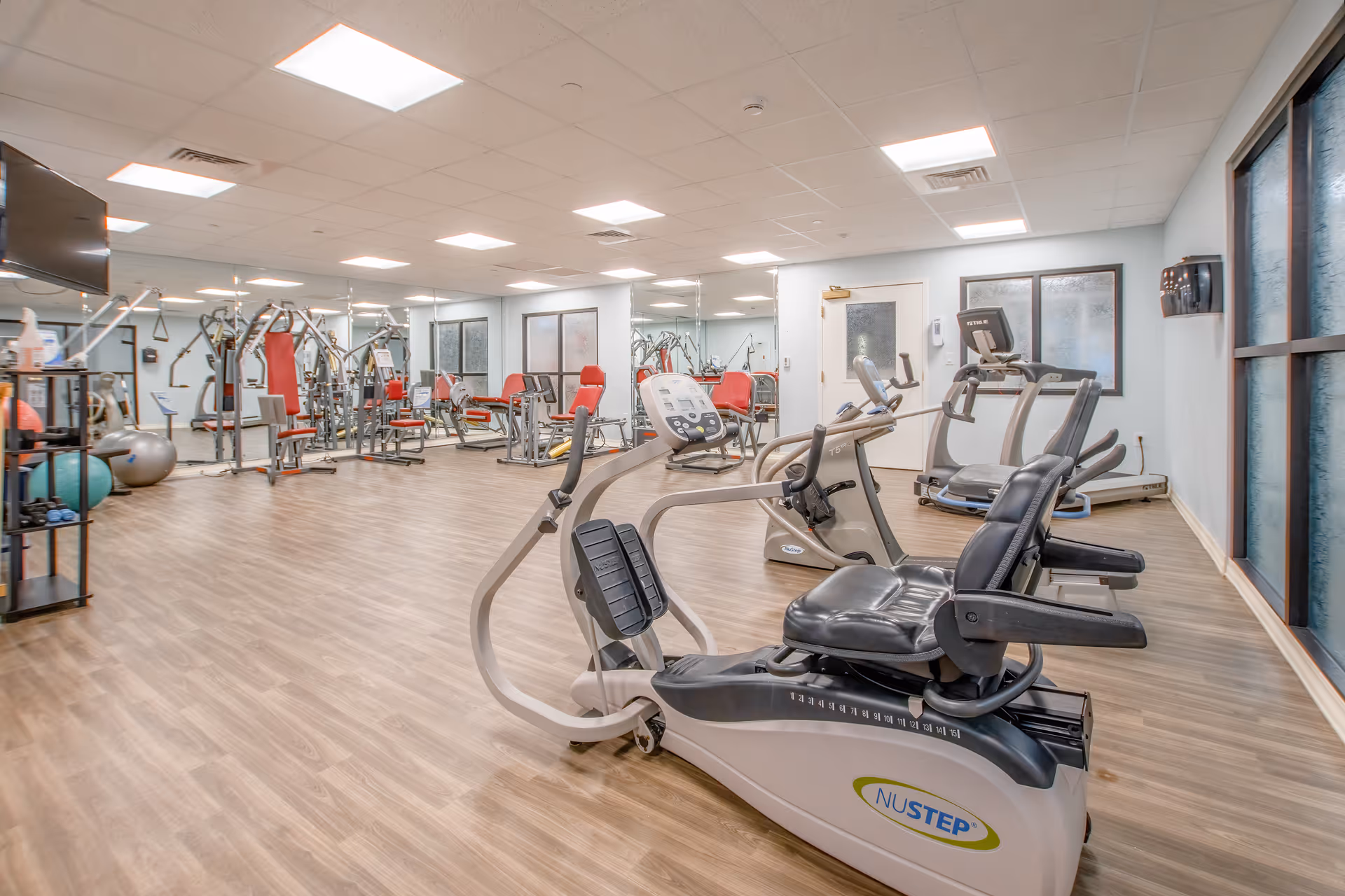 A spacious fitness room with various exercise equipment including recumbent bikes, treadmills, weight machines, and exercise balls. The room has wood-style flooring, large mirrors on one wall, and several windows allowing natural light. The ceiling has recessed lighting, and the walls are painted light blue.