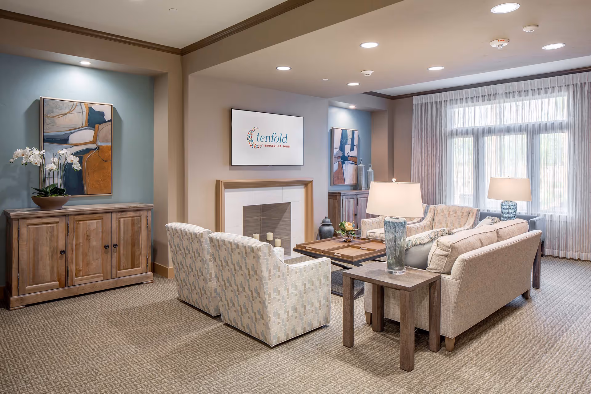 A cozy living room area with beige and patterned armchairs, a beige sofa, wooden side tables with lamps, a wooden coffee table, and a fireplace with a mounted TV above it displaying the Tenfold Bruceville Point logo. The room has large windows with sheer white curtains, a wooden cabinet with a potted orchid and abstract artwork on the wall.