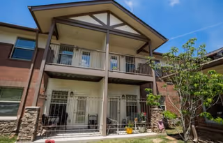 Exterior view of a two-story residential building with balconies on the upper floor and patios on the ground floor, surrounded by a small grassy area and a tree under a clear blue sky.