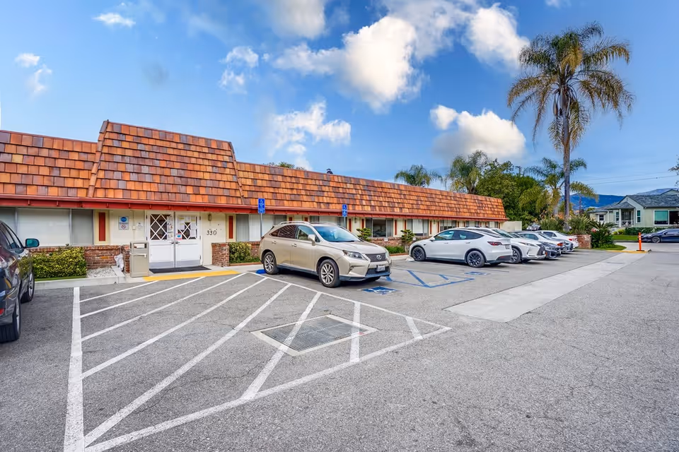 Exterior view of Leisure Glen Care Center showing a single-story building with a brown tiled roof, several parked cars in front, a clear blue sky with some clouds, and palm trees in the background.
