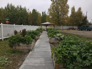 A paved walkway flanked by raised garden beds filled with green plants leads to a wooden gazebo. The area is bordered by a white fence on the left and a parking lot with a green vehicle on the right. Trees with autumn foliage are visible in the background under a cloudy sky.