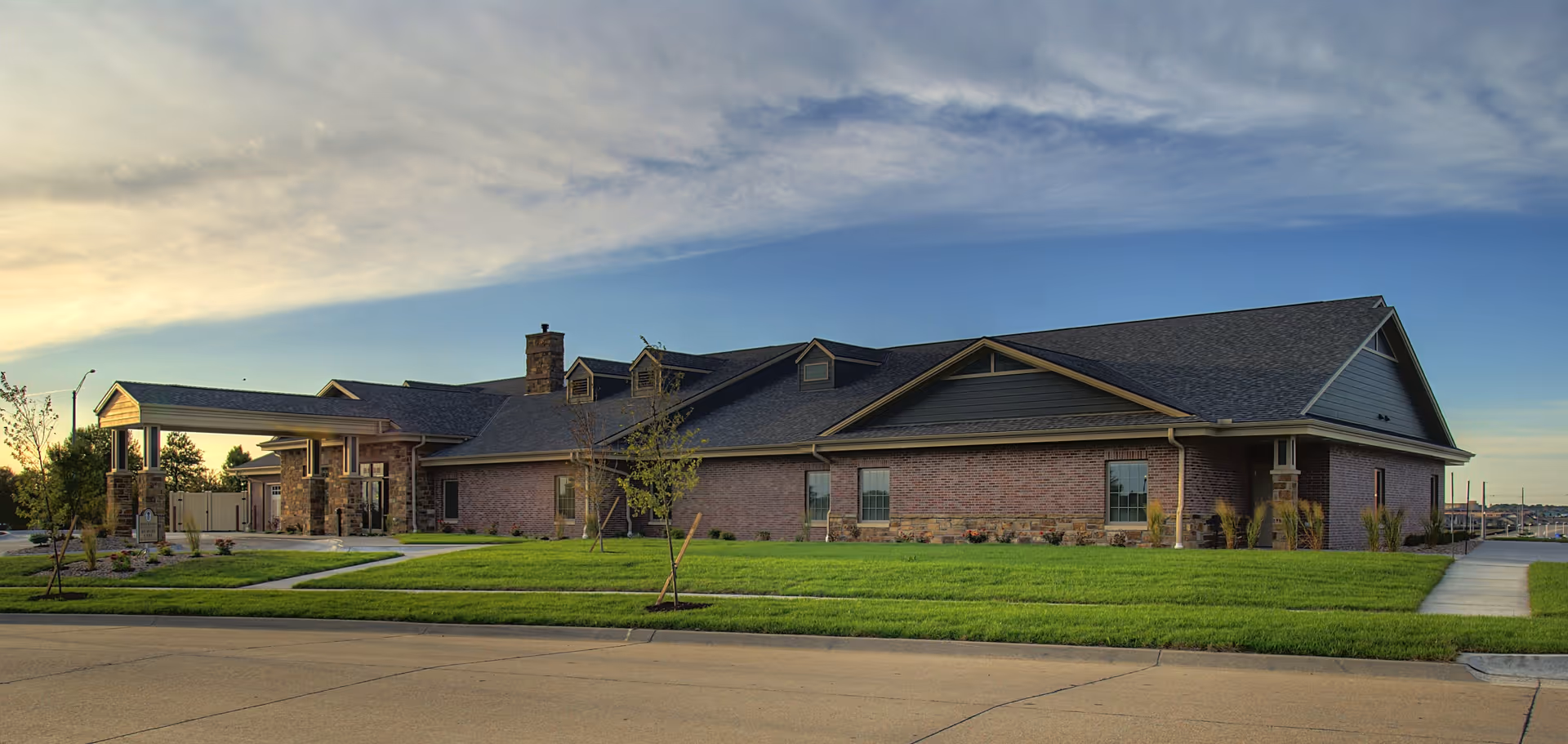 Exterior view of a single-story senior living facility building with brick walls, a covered entrance, and a well-maintained lawn under a partly cloudy sky during sunset.