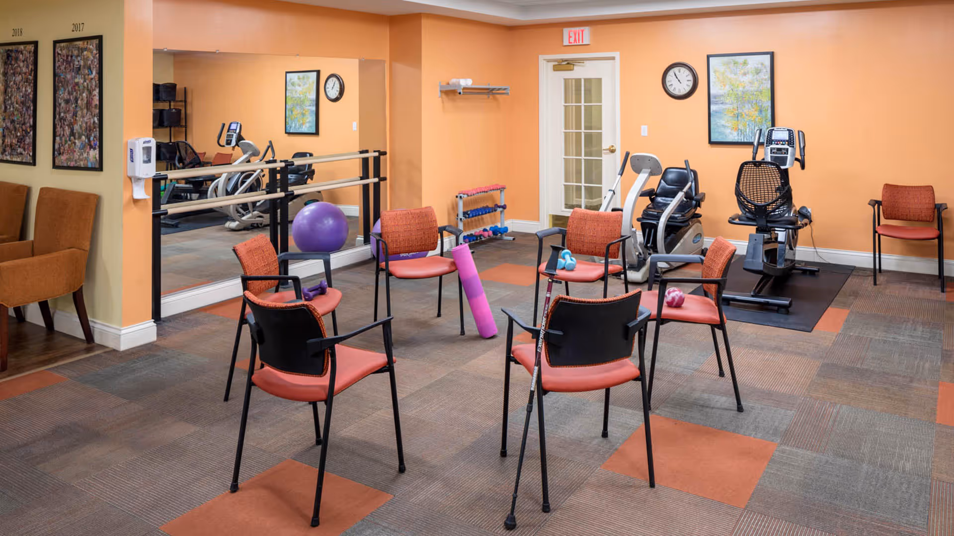A fitness room in a senior living facility with six orange chairs arranged in a circle, some holding small exercise weights and a purple foam roller. The room has exercise equipment including a recumbent bike and elliptical machine, a large mirror on one wall, a purple exercise ball, and a rack of small dumbbells. The walls are painted orange and beige, with framed artwork and a clock. There is a door with a glass panel and an exit sign above it.