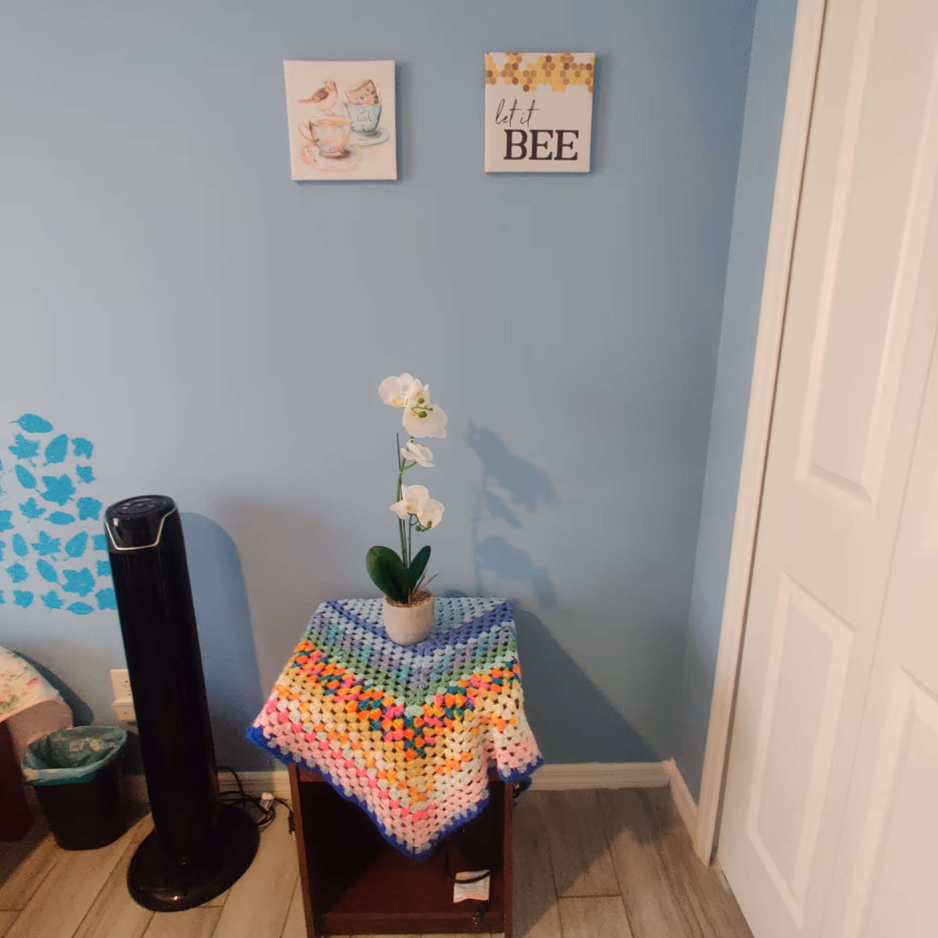 A corner of a room with light blue walls featuring two small wall decorations, one with birds and teacups and the other with the text 'let it BEE'. Below the decorations is a small wooden table covered with a colorful crocheted blanket and a white orchid plant in a pot. To the left of the table is a black tower fan and a small trash bin. On the right side, there is a white closet door.
