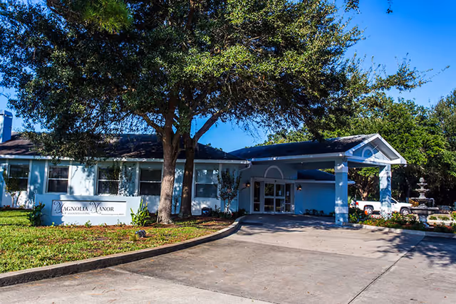 Exterior view of Magnolia Manor Assisted Living facility showing a single-story building with a covered entrance, surrounded by trees and greenery under a clear blue sky. A sign with the facility's name is visible on the lawn near the driveway.