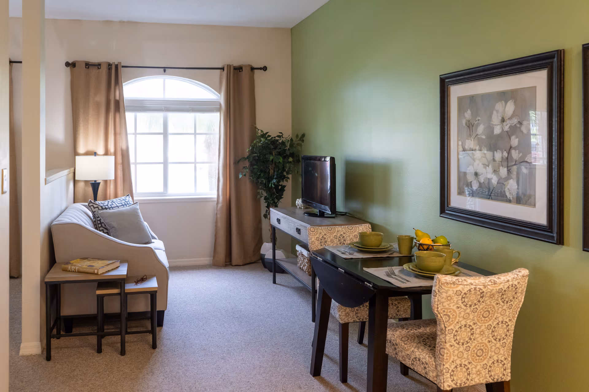 A cozy living and dining area with a beige sofa next to a window with tan curtains. A small side table with books and glasses is beside the sofa. Across from the sofa is a TV on a console table, and a dining table set for two with green dishes and patterned chairs. The walls are painted green and decorated with framed floral artwork.