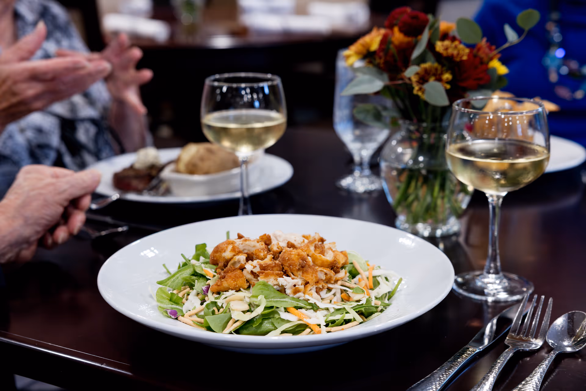 A close-up of a dining table with a plate of salad topped with fried chicken pieces, two glasses of white wine, and a vase with a bouquet of flowers. In the background, there are blurred images of people dining and another plate with a baked potato and steak.