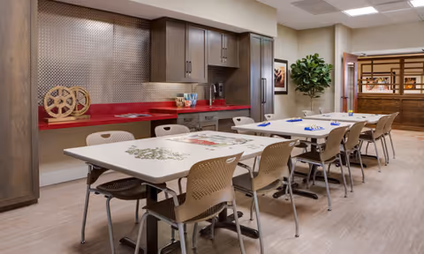 Communal interior room with rows of tables and chairs and a kitchenette with red countertops and cabinets along the back wall.