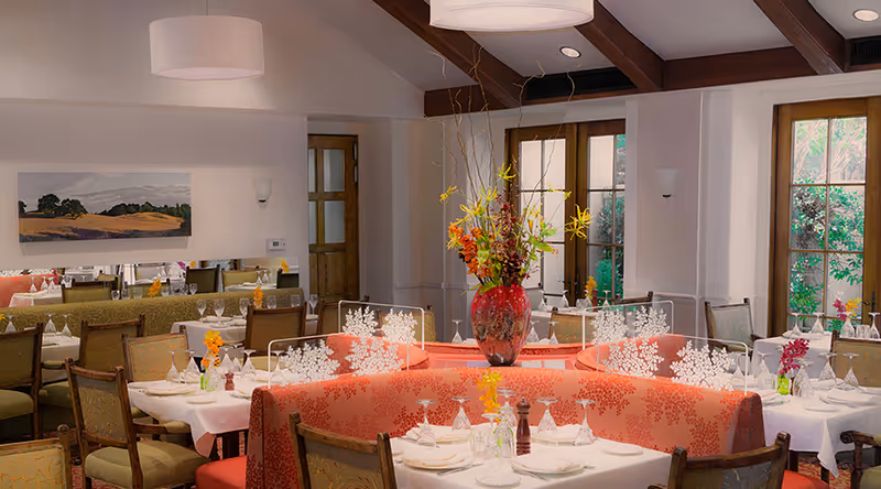 A well-lit dining room with round tables set with white tablecloths, glassware, and napkins. The room features a large floral centerpiece in a red vase on a circular booth with red patterned upholstery. There are wooden chairs with green cushions around the tables, large windows letting in natural light, and a landscape painting on the wall.