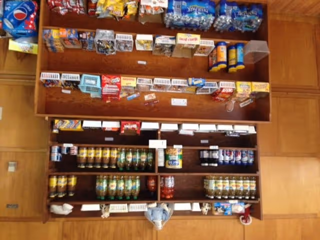 Overhead view of a wooden snack and beverage display shelf stocked with various snacks, chips, bottled water, soda cans, and other drinks in a retail or community setting.