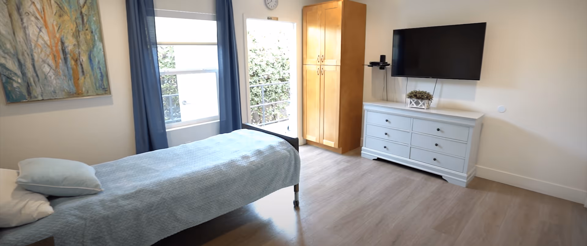Bright single-occupancy bedroom with a bed, dresser and wall-mounted TV next to windows and a door.