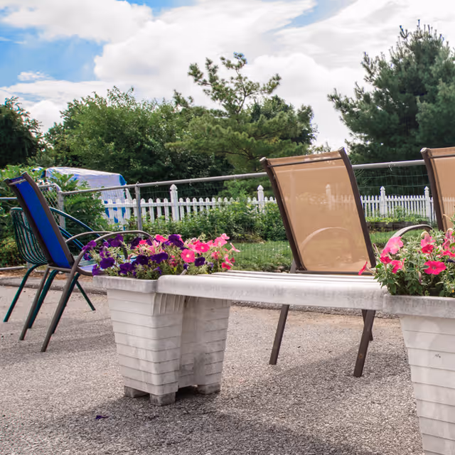 Outdoor patio with chairs and a concrete bench planter holding pink and purple flowers, with a white picket fence and trees in the background.