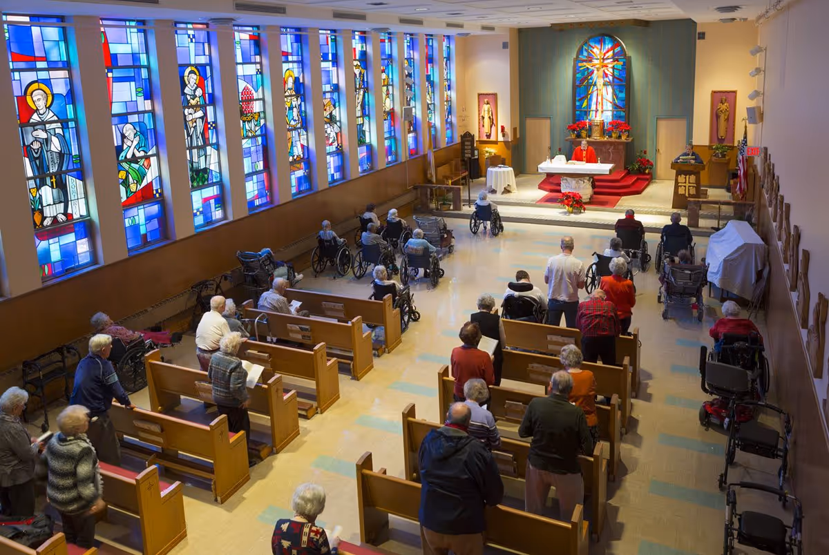 Interior view of a chapel with stained glass windows along one wall. Several elderly people, some in wheelchairs, are seated or standing in wooden pews facing an altar where a priest is conducting a service.