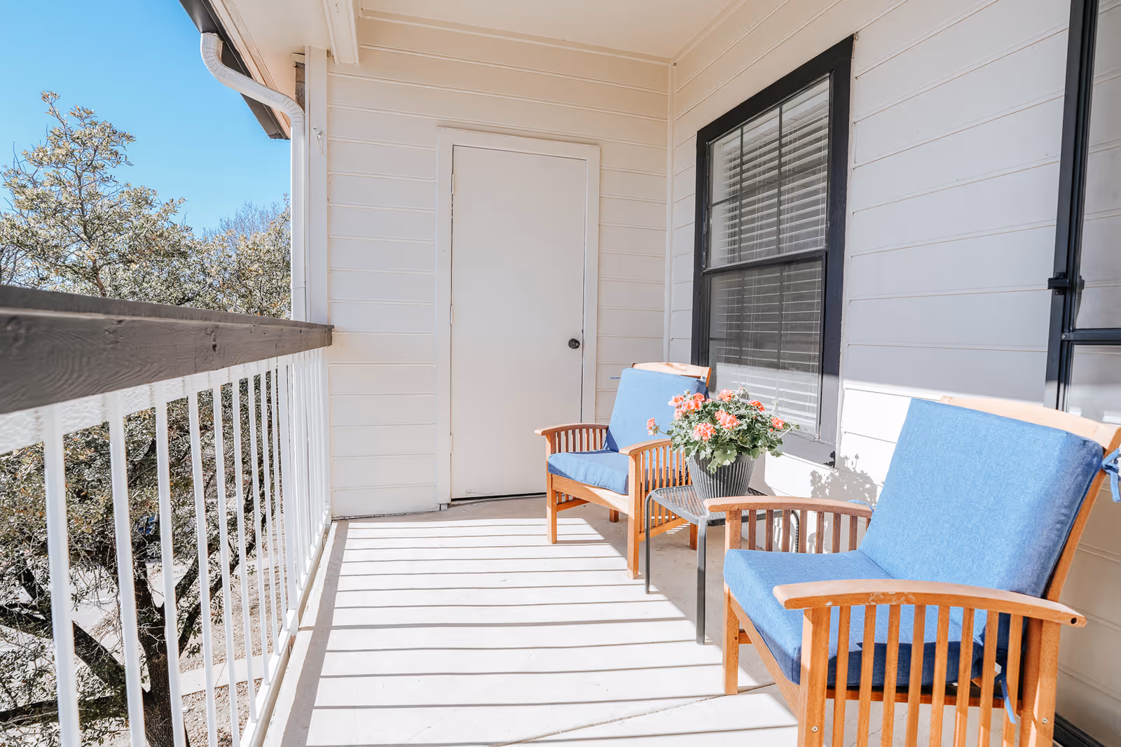 A sunny balcony with two wooden chairs with blue cushions, a small side table holding a potted flowering plant, and a white exterior wall with a door and window.