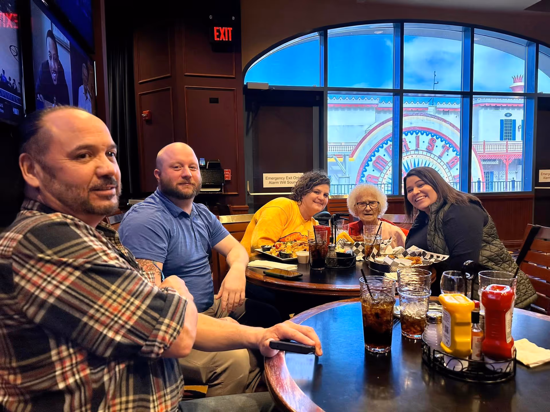 A group of five people sitting around a table in a restaurant, enjoying a meal together. The group includes an elderly woman and four adults, with plates of food and drinks on the table. Large windows in the background show a colorful exterior sign.