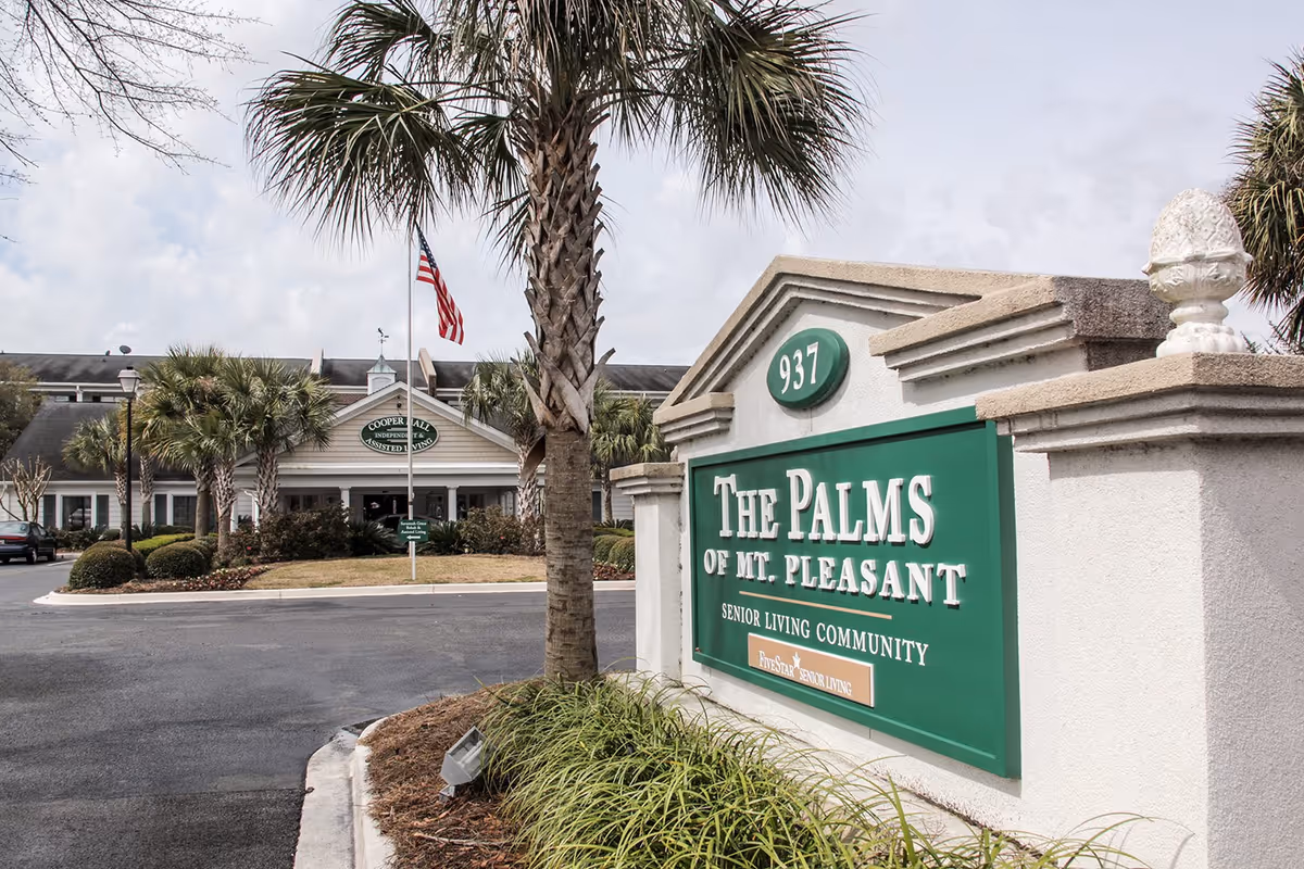Entrance sign for The Palms of Mt. Pleasant senior living community with palm trees, a driveway, and the facility building in the background under a cloudy sky.