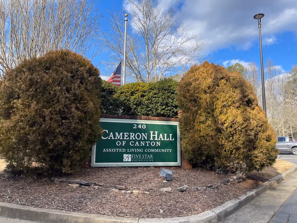 Entrance sign reading "240 Cameron Hall of Canton Assisted Living Community" surrounded by shrubs with an American flag in the background.