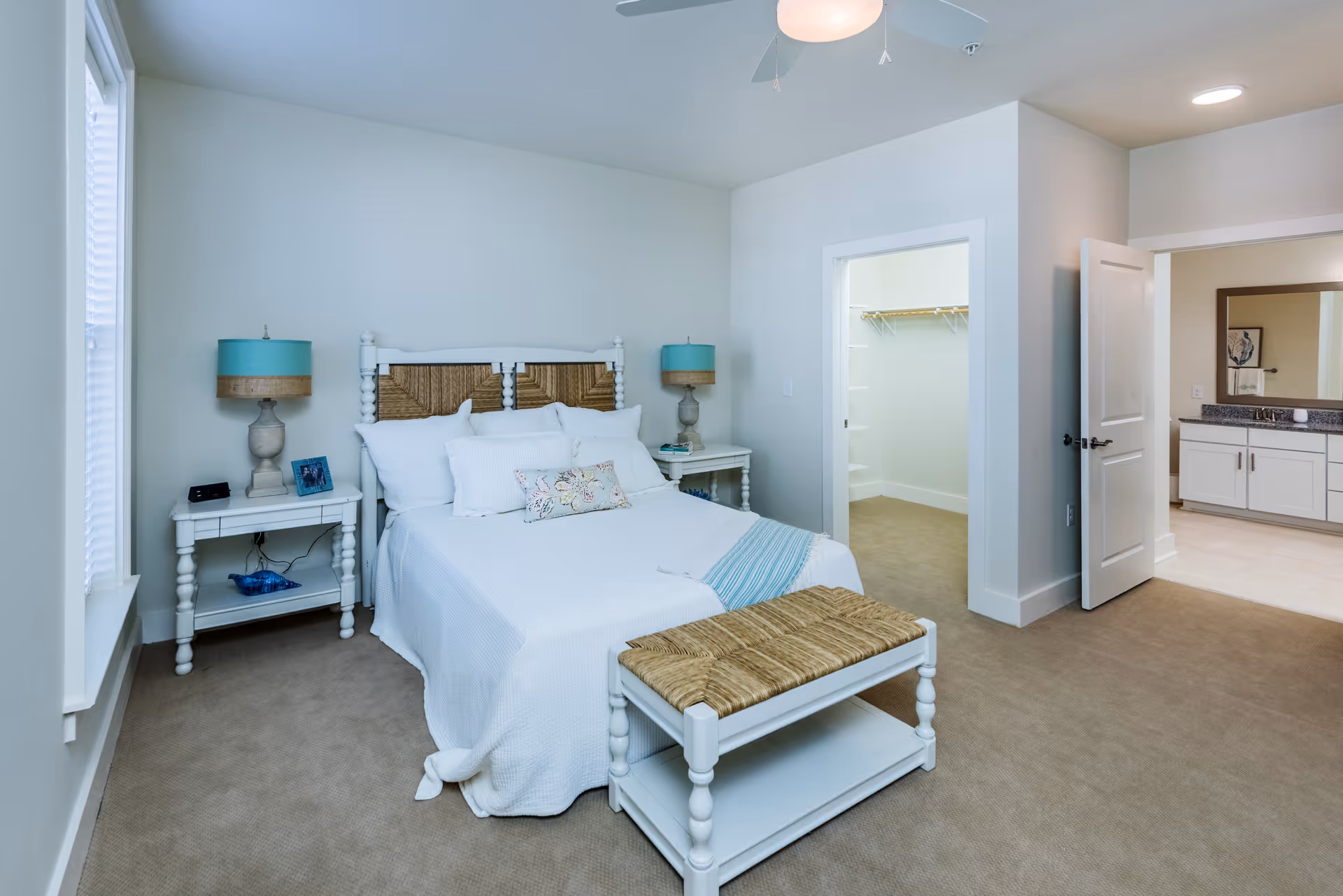 A neatly made bed with white bedding and multiple pillows in a bedroom. The bed has a white frame with a woven headboard and a matching bench at the foot. There are two white nightstands with turquoise lamps on either side of the bed. To the right, there is an open door leading to a walk-in closet and another door leading to a bathroom with a large mirror and white cabinetry. The room has beige carpeting and light-colored walls.