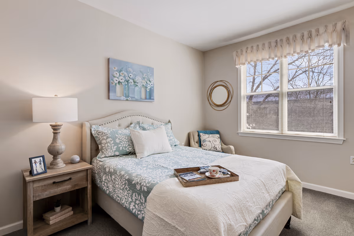 A cozy bedroom with a bed featuring a light blue and white floral patterned comforter and pillows. A wooden nightstand with a lamp, a small framed photo, and a decorative item is beside the bed. Above the bed hangs a painting of flowers in vases. There is a window with blinds and a valance, and a cushioned chair with a decorative pillow is placed near the window. A round mirror with a gold frame is mounted on the wall next to the window. A tray with a cup and a book is on the bed.