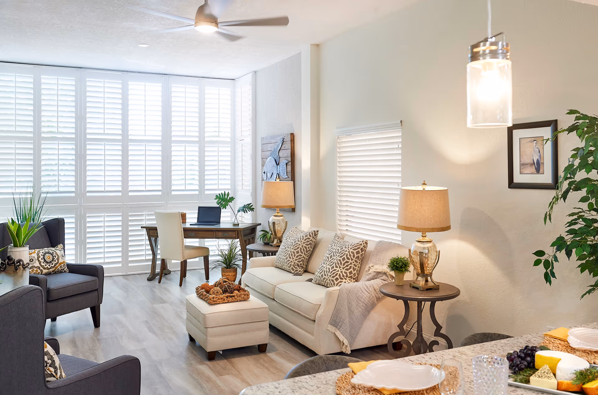 A bright and cozy living room with light-colored walls and flooring. The room features a beige sofa with patterned pillows and a matching ottoman, two dark gray armchairs with decorative pillows, a wooden desk with a chair and a laptop near large windows with white plantation shutters. There are two side tables with lamps, a hanging pendant light, a framed bird picture on the wall, and various green plants adding a touch of nature. In the foreground, a dining counter is set with plates, glasses, and a cheese platter.