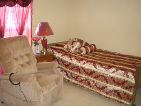 A cozy room with a single bed covered in a red and gold patterned bedspread with matching pillows. Next to the bed is a wooden nightstand with a red lamp, a small bouquet of pink flowers, and a framed photo. A beige recliner chair is positioned nearby. The window has pink curtains with a decorative valance.