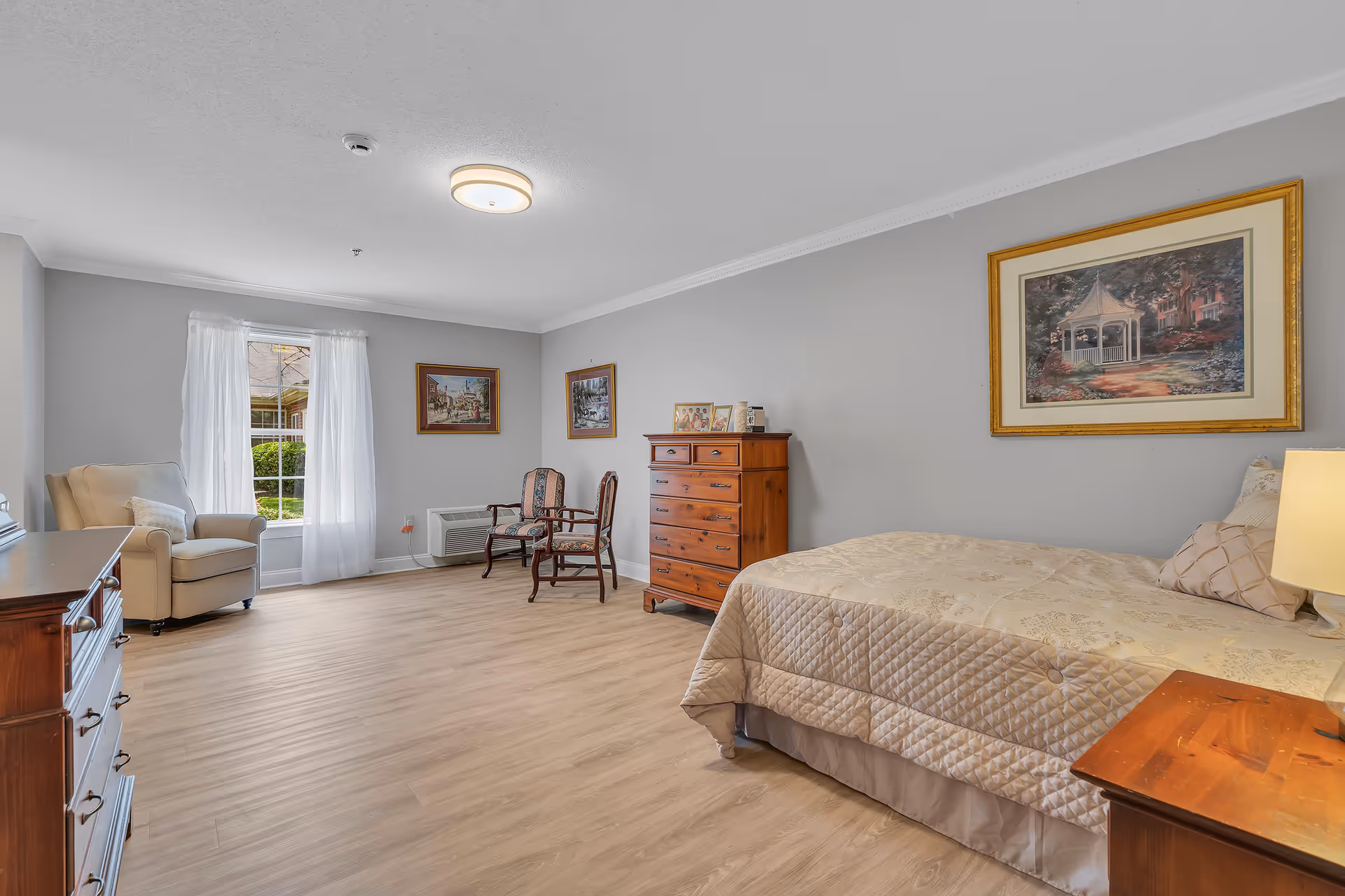 A spacious bedroom with light wood flooring, a bed with a beige quilt, a wooden dresser, two chairs, an armchair near a window with white curtains, and framed artwork on the light gray walls.