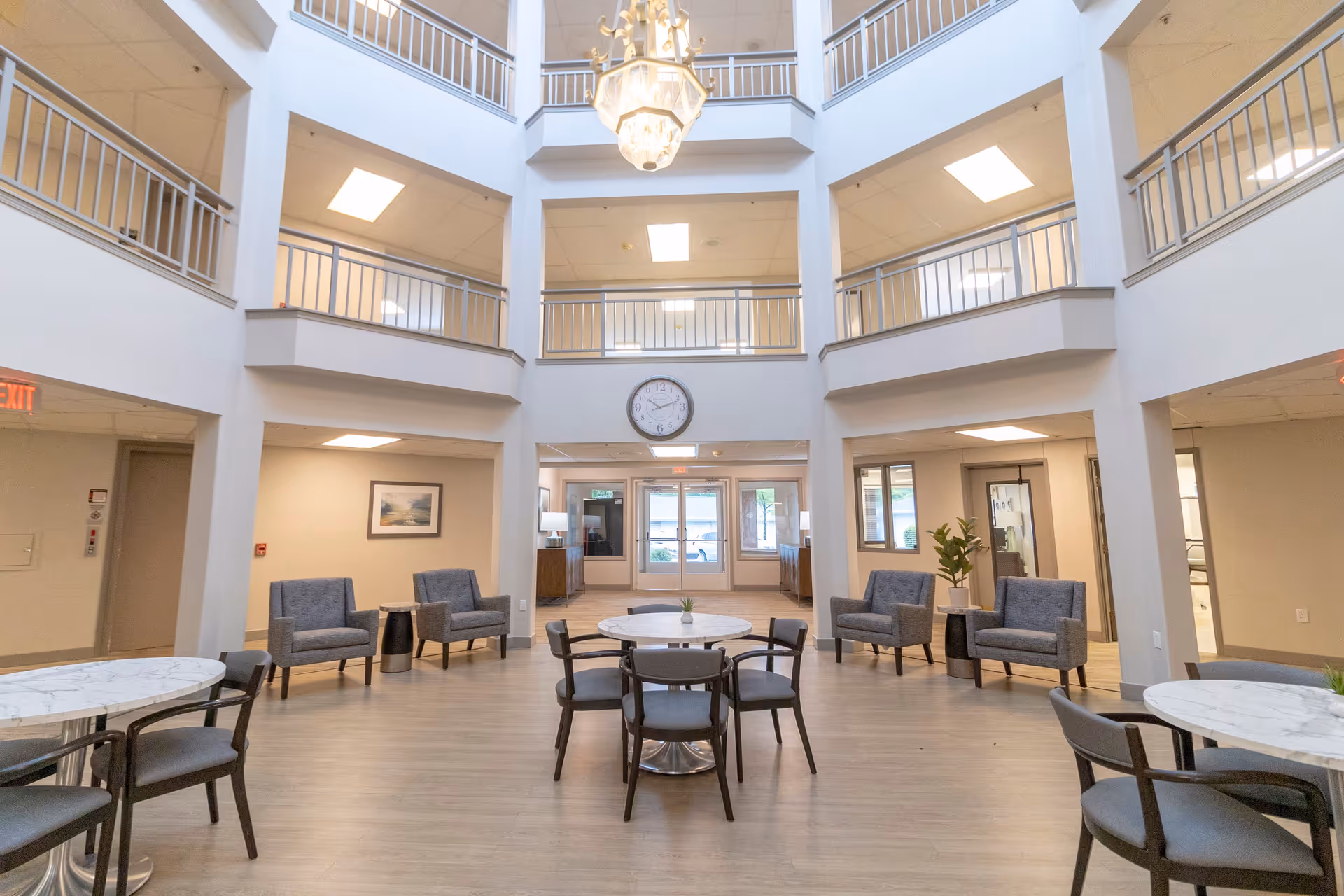 A spacious, well-lit common area inside a senior living facility with multiple seating arrangements including round tables with chairs and cushioned armchairs. The room features a high ceiling with a large chandelier, a clock on the wall, and balconies on the upper floors overlooking the area. The flooring is light wood, and there are large windows and doors at the far end letting in natural light.
