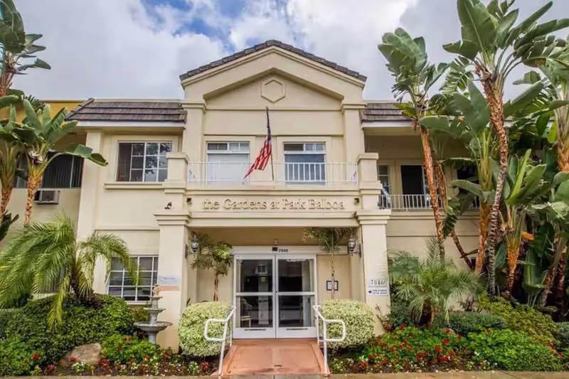 Front exterior view of The Gardens at Park Balboa building with beige walls, a tiled roof, an American flag above the entrance, and surrounded by lush greenery and palm trees.