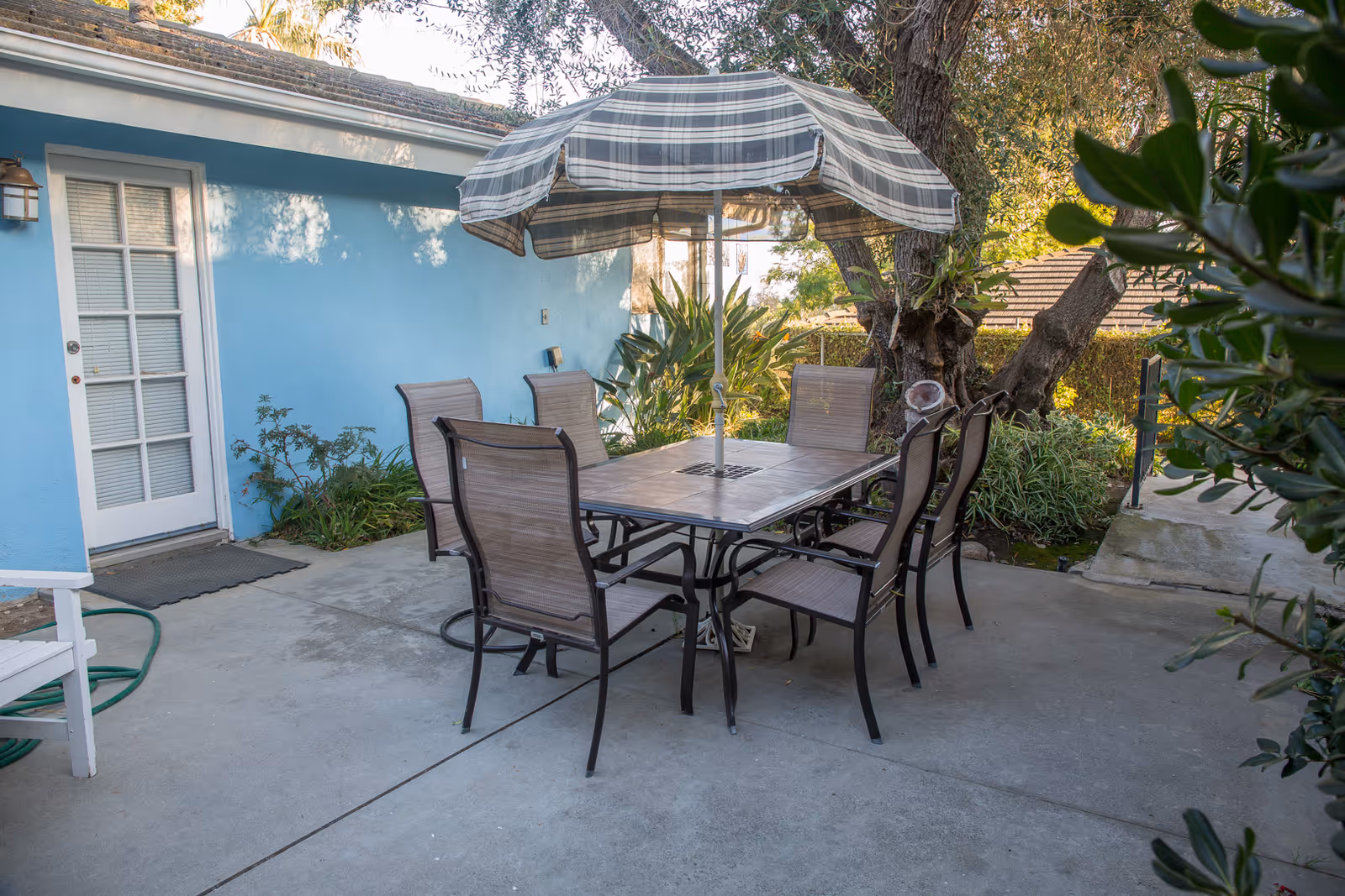 Outdoor patio area with a rectangular table surrounded by six chairs. A large striped umbrella is positioned in the center of the table. The patio is adjacent to a blue building with a white door and windows. There are plants and trees around the patio providing shade and greenery.