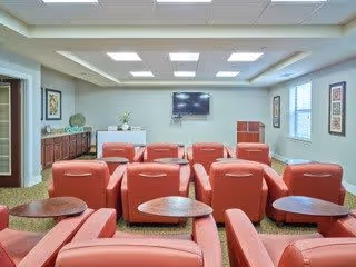A bright room with multiple red leather recliner chairs arranged in rows facing a wall-mounted flat screen TV. The room has a light-colored ceiling with recessed lighting, beige walls with framed artwork, and windows letting in natural light. There is a wooden cabinet along one wall with decorative items on top.
