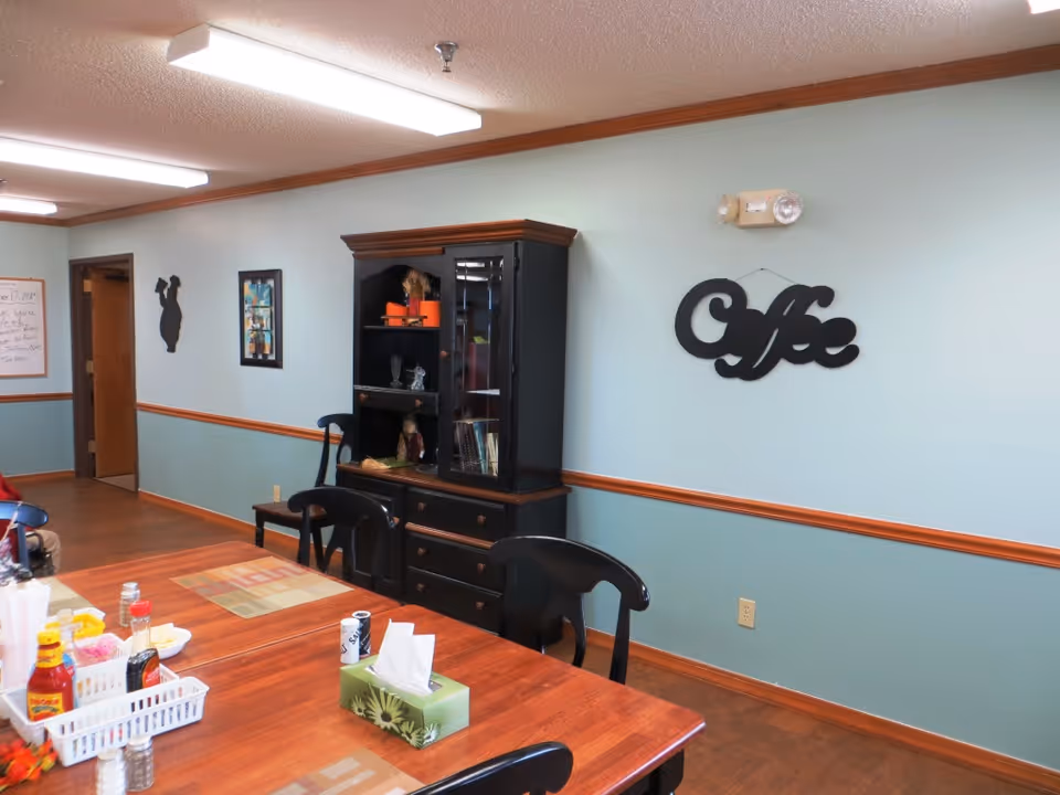 A dining area with a wooden table and black chairs. On the table are condiments, napkins, and placemats. Against the wall is a black cabinet with shelves holding decorative items. The wall is painted light blue with a wooden trim, and a decorative sign that reads 'Coffee' is hanging on the wall. There is a doorway and framed artwork on the far wall.