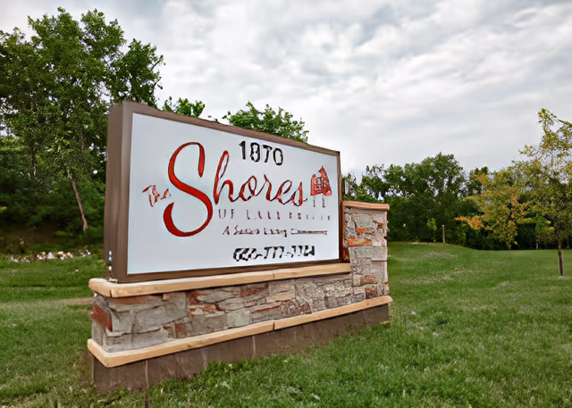 Stone monument sign reading 'The Shores of Lake Phalen' on a grassy lawn with trees and a cloudy sky.