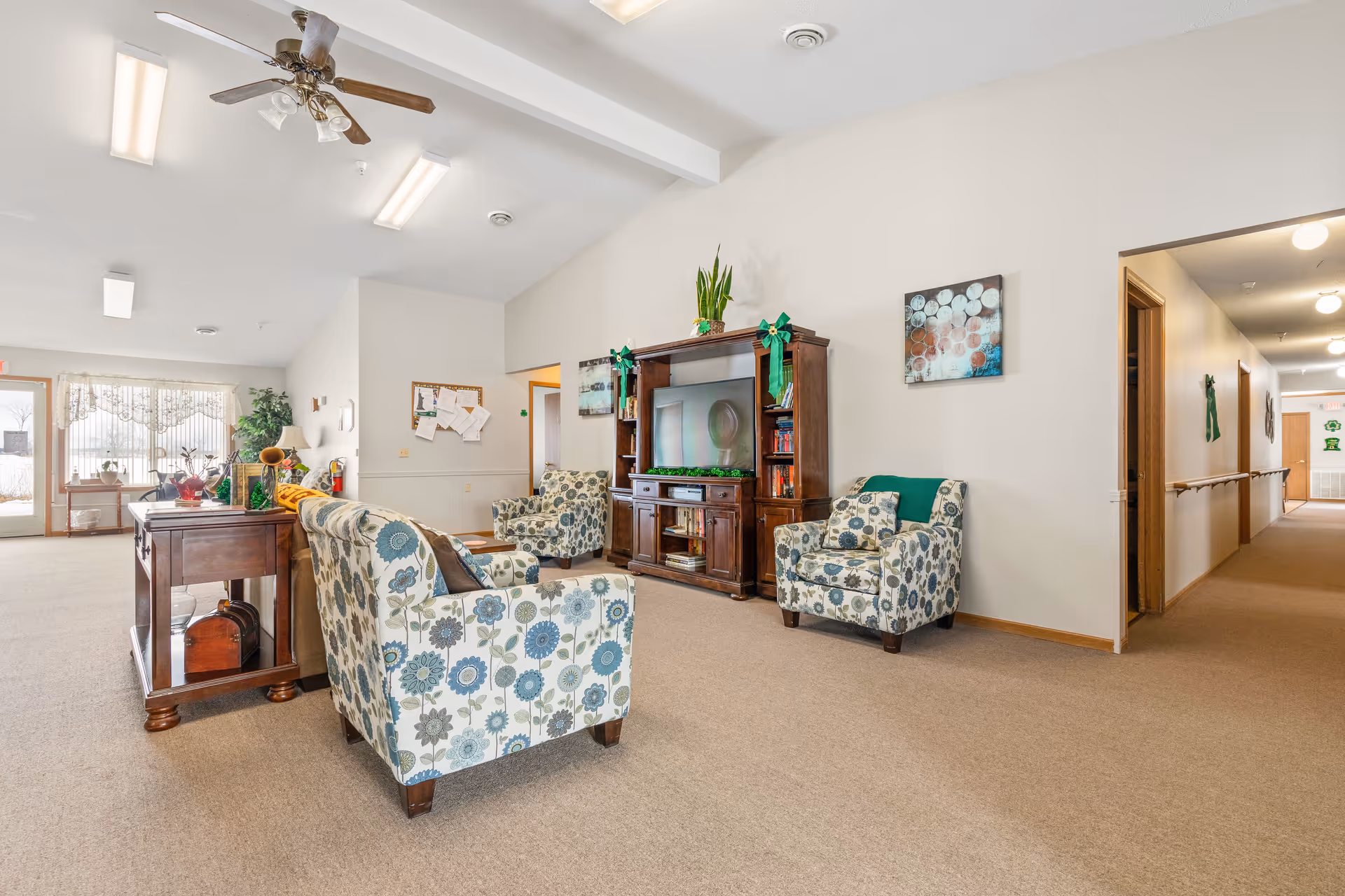 A bright and spacious living room area in a senior living facility with floral patterned armchairs, a wooden entertainment center with a TV, and a wooden table with decorative items. There is a hallway to the right and large windows with sheer curtains in the background letting in natural light.