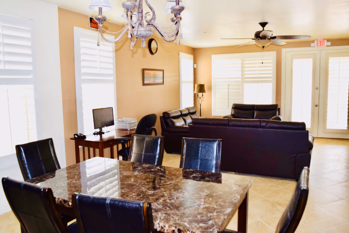 A bright living area with a marble dining table surrounded by six black leather chairs in the foreground. In the background, there are two dark leather sofas, a ceiling fan, a floor lamp, a small desk with a computer monitor and chair, and several windows with white plantation shutters. The walls are painted beige, and there is a chandelier hanging above the dining table.