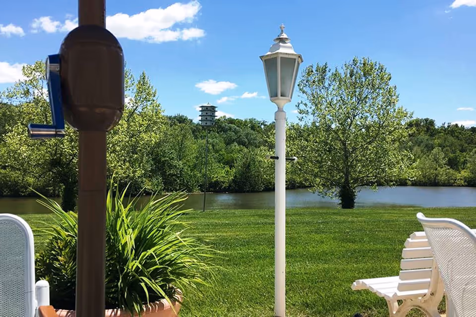 Patio view overlooking a grassy lawn and river with trees, a white lamp post, potted plants, and a white bench under a blue sky.
