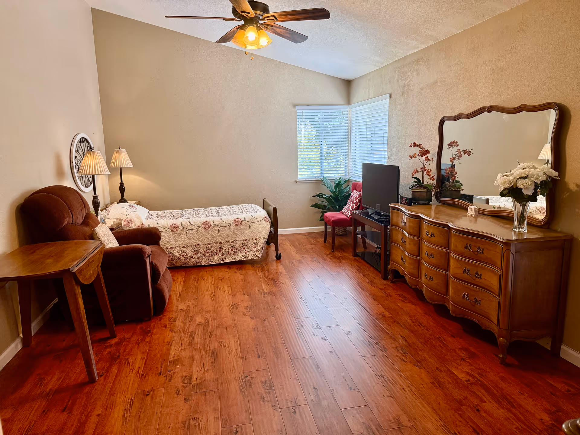 A senior living bedroom with a single bed covered in floral bedding, a brown recliner chair, a wooden side table, a dresser with a large mirror, a TV on a stand, a red chair, and a window with white blinds. The room has wooden flooring, beige walls, and a ceiling fan with lights.