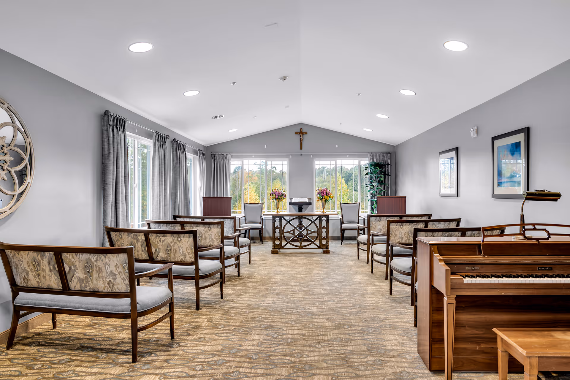 A bright, spacious room with rows of wooden chairs with patterned cushions arranged facing a wooden table with floral arrangements and a lectern. Large windows with gray curtains let in natural light, and a piano is positioned on the right side of the room. The walls are painted gray and decorated with framed artwork and a decorative wall piece. A crucifix hangs on the wall above the table at the far end of the room.