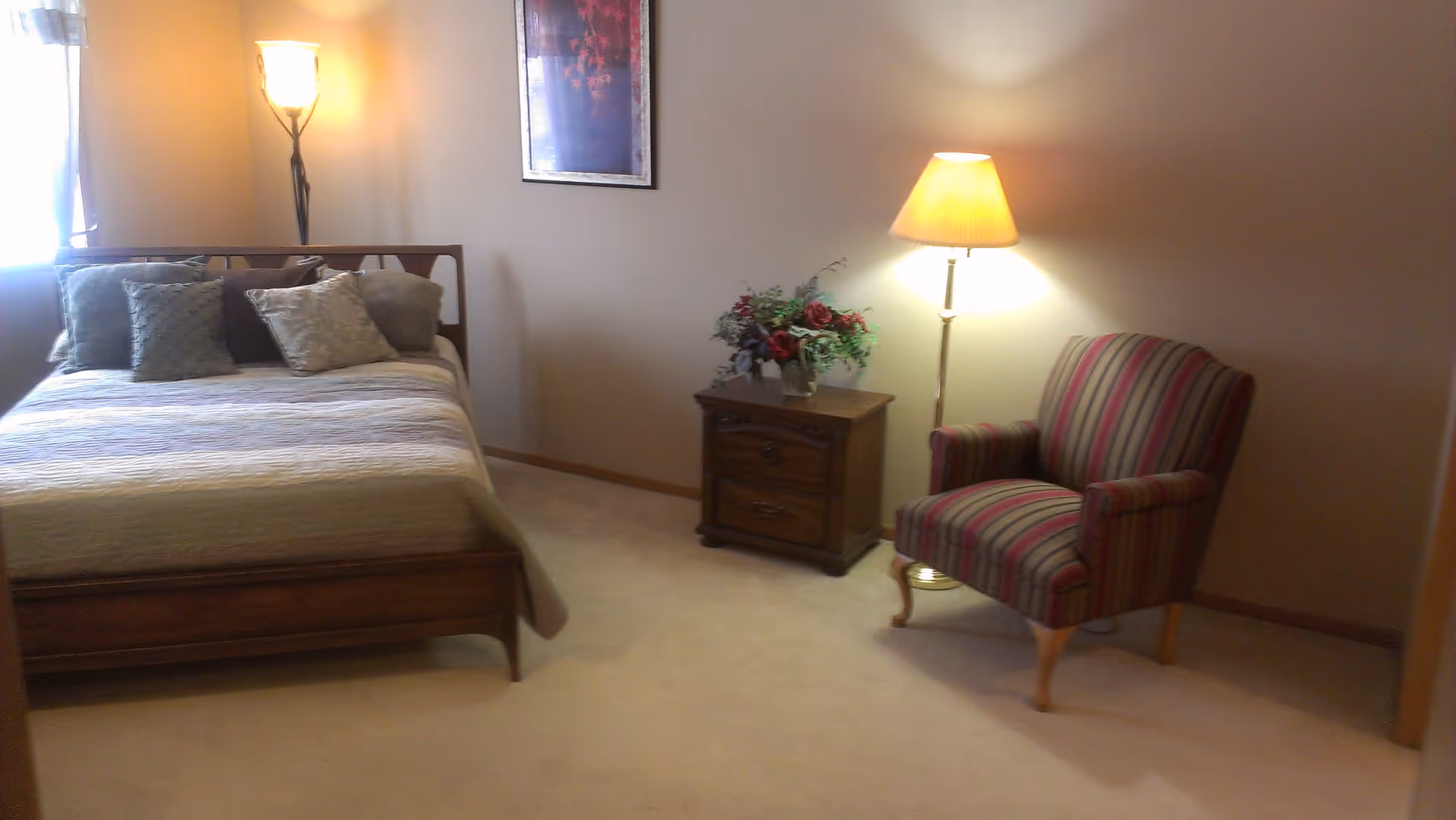 A bedroom with a wooden bed frame and a neatly made bed with multiple pillows. There is a wooden nightstand with a floral arrangement and a floor lamp with a yellow shade next to a striped upholstered armchair. A framed picture hangs on the wall above the nightstand, and a tall lamp is positioned in the corner near a window with curtains.