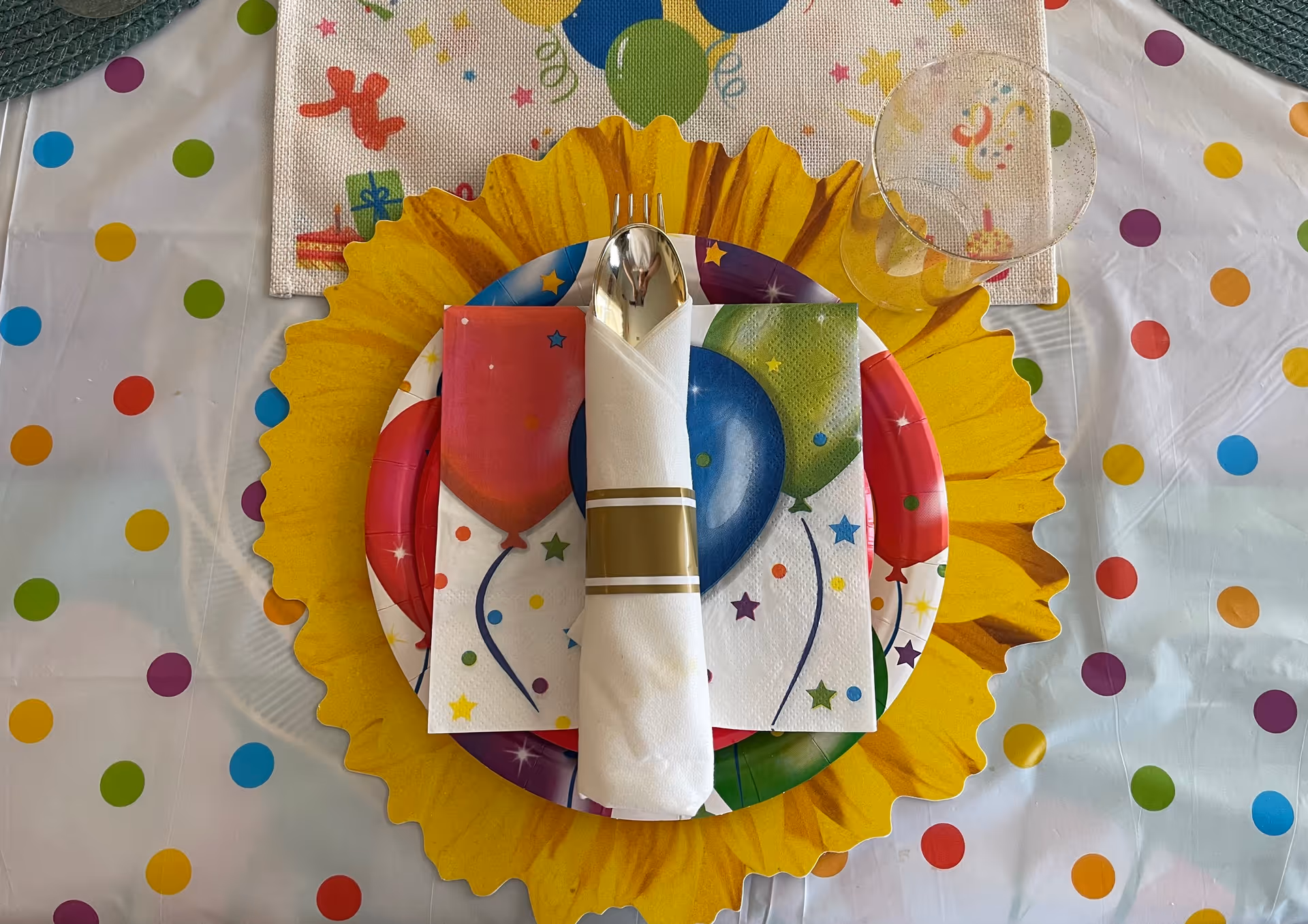 A colorful party table setting with a sunflower-shaped yellow placemat, a plate with balloon-themed napkins, a rolled white napkin with a gold band holding a spoon and fork, and a clear plastic cup on a polka dot tablecloth.