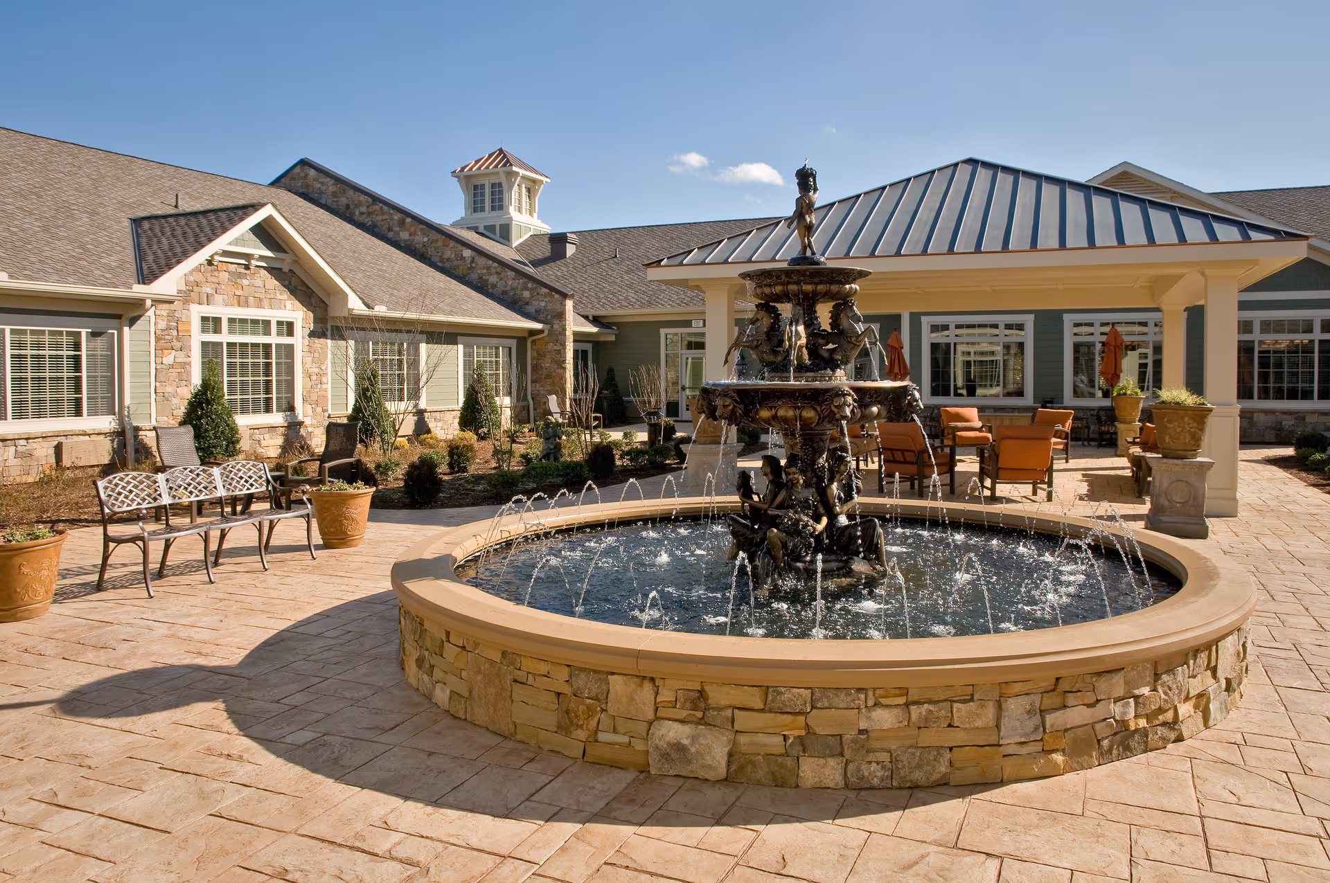 Outdoor courtyard area of a senior living facility featuring a large decorative stone fountain with water jets, surrounded by a paved patio with benches, potted plants, and outdoor seating under a covered area. The building exterior has stone and siding with multiple windows and a cupola on the roof under a clear blue sky.