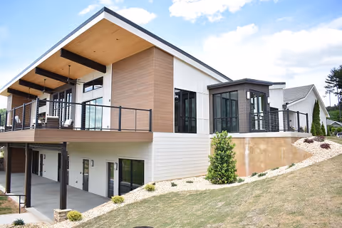 Modern two-story building with a covered upper deck, large windows, and sloped landscaping.