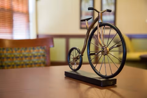 A decorative metal sculpture of a vintage bicycle placed on a wooden table in a room with patterned chairs and a window with blinds in the background.