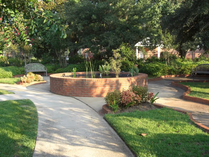 A peaceful outdoor garden area with a circular brick planter filled with plants in the center, surrounded by curved concrete pathways and green grass. There are wooden benches placed along the pathways and large trees providing shade in the background.