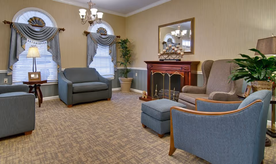 A cozy living room with beige walls and carpeted floor, featuring two arched windows with decorative curtains, a chandelier, a wooden fireplace with a mirror above it, a gray armchair with matching ottoman, a loveseat, a side table with a lamp and picture frame, and several potted plants.